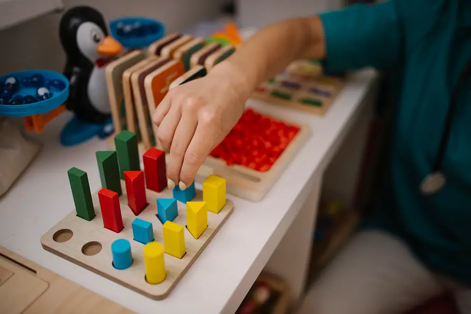 Ahşap tabla üzerinde renkli geometrik blokları yerleştiren bir el; arka planda başka eğitim materyalleri