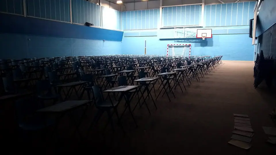 Loş bir spor salonunda sıra sıra dizilmiş boş sınav masaları