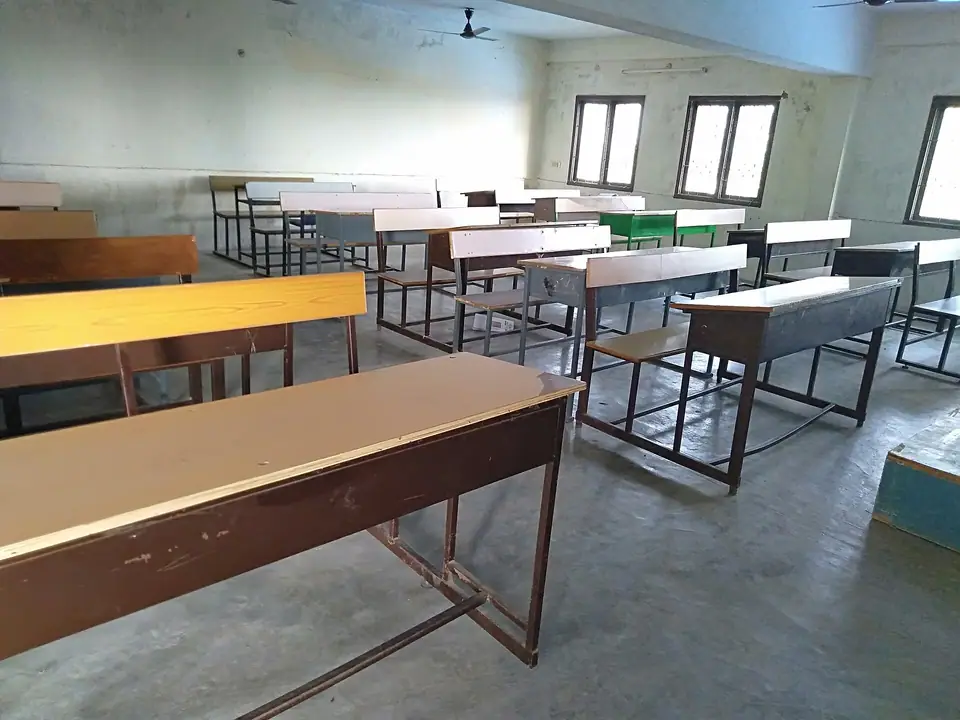 Pencereli bir derslikte sıra sıra dizilmiş boş ahşap ve metal sıralar