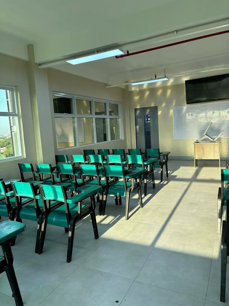 Güneş alan boş bir derslikte sıra sıra yeşil sandalyeler ve beyaz tahta