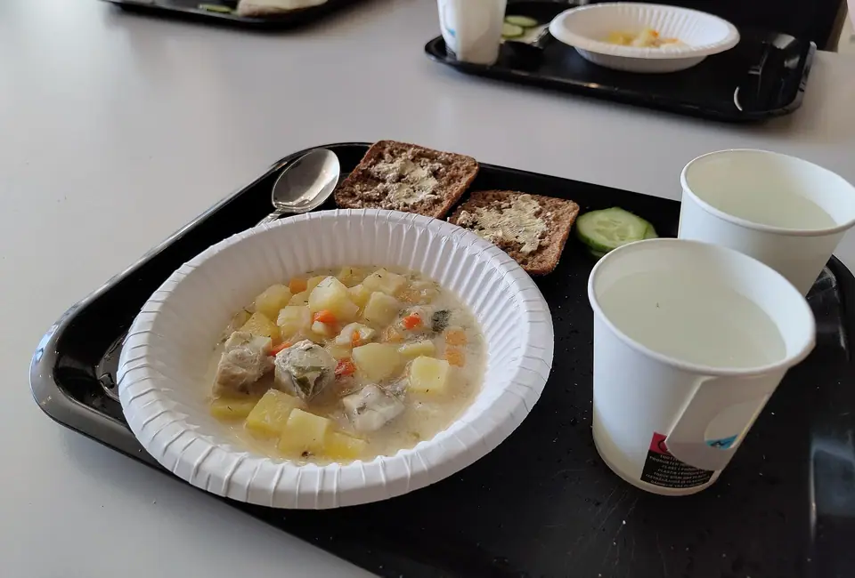 Siyah tepside çorba kâsesi, üzerine sürülmüş iki dilim tam tahıllı ekmek, salatalık ve iki kâğıt bardak
